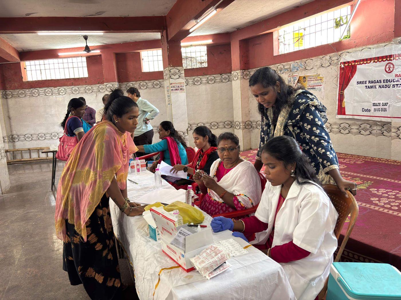 Health workers in white coats screening people at a camp table